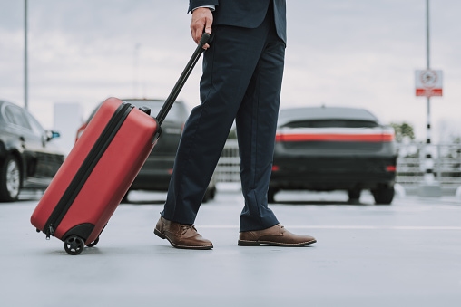 Cropped portrait of guy wearing black costume with suitcase staying on the parking lot with cars on background