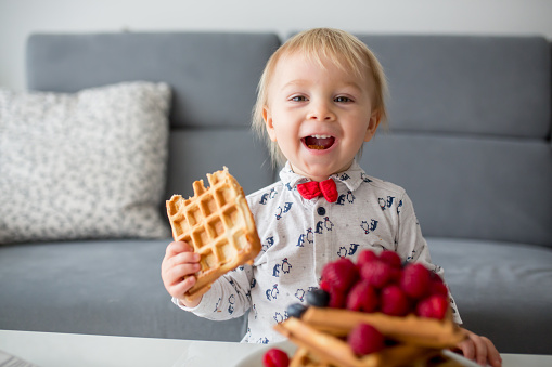 Sweet toddler birthday boy, eating belgian waffle with raspberries, blueberries, cocnut and chocolate at home