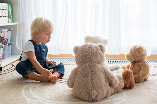 Little toddler boy, reading a book to his teddy bear friends at home, sitting in living room