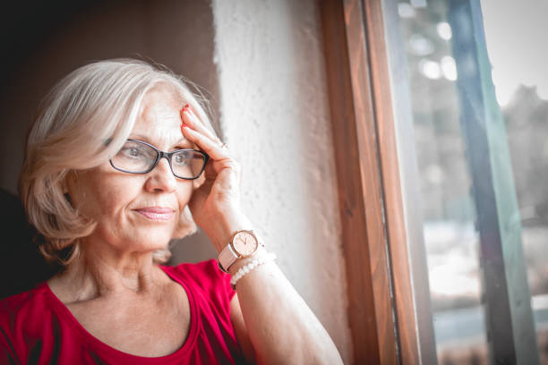 mujer solitaria de la tercera edad - pensive senior adult looking through window indoors fotografías e imágenes de stock