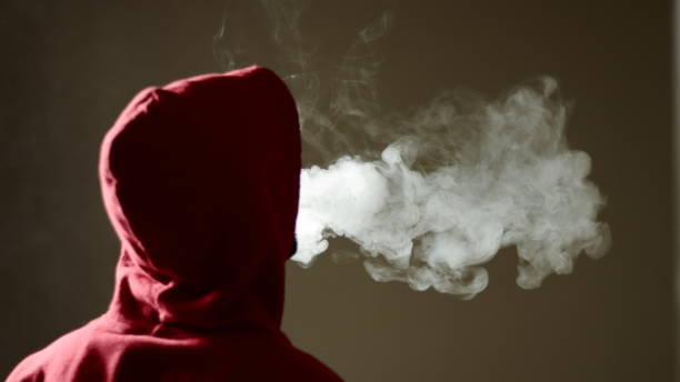 赤いパーカーを着た若い男性が喫煙し、厚い蒸気を吐き出し、孤立した後方の景色を呼び起こします - 電子タバコ ストックフォトと画像