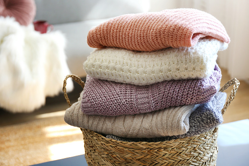 Bunch of stacked knitted pastel color sweaters with different knitting patterns perfectly folded in wicker basket on table, living room background. Fall winter season knitwear. Close up, copy space.
