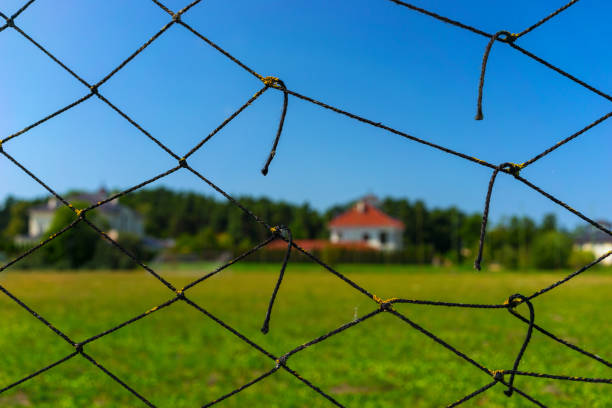 A house with a red roof under a blue sky near a forest next to a green field through an old torn rope mesh