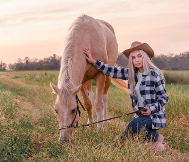 junge blonde mädchen in einem cowboyhut und blaue jeans mit einem beige pferd auf der ranch gekleidet - cowboy blue meadow horizontal stock-fotos und bilder