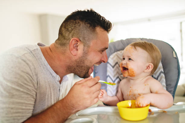 小さな赤ちゃんが彼女の夕食を食べて、側に父親と混乱を作る - feeding ストックフォトと画像