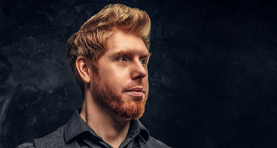 Close-up portrait of a handsome redhead man with stylish hair and beard in studio against a dark textured wall