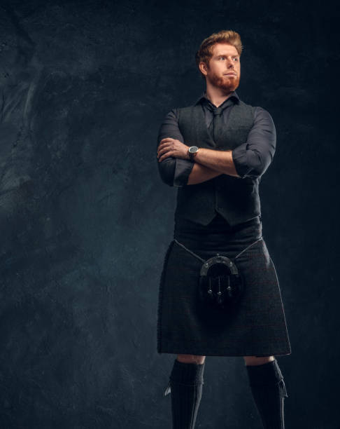 Redhead man dressed in an elegant vest with tie and kilt in studio against a dark textured wall Handsome redhead man dressed in an elegant vest with tie and kilt posing with his arms crossed in studio against a dark textured wall kilt stock pictures, royalty-free photos & images