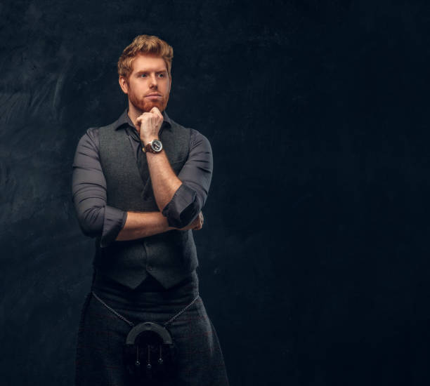 istock Redhead man dressed in an elegant vest with tie and kilt posing with thoughtful look in studio against a dark textured wall 1175932932