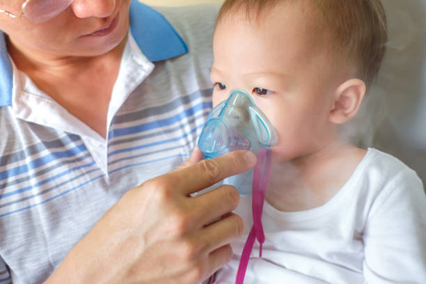 Sick little kid with respiratory problem with oxygen mask breathes through nebulizer at doctor clinic Asian father helping his toddler son with inhalation therapy by the mask of inhaler. Sick little kid with respiratory problem with oxygen mask breathes through nebulizer at doctor clinic / hospital pediatric nebulizer mask stock pictures, royalty-free photos & images