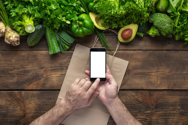istock Order food online Man holds smartphone with blank screen for your text message or design on wooden background with fresh green vegetables 1175963063