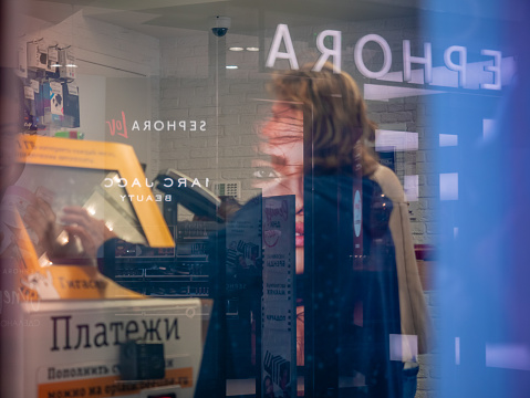 Moscow, Russia - September 14, 2019: Advertising reflections of Sephora store in glass of shopping center window. Woman makes payment for mobile phone on self-service checkout counter Beeline
