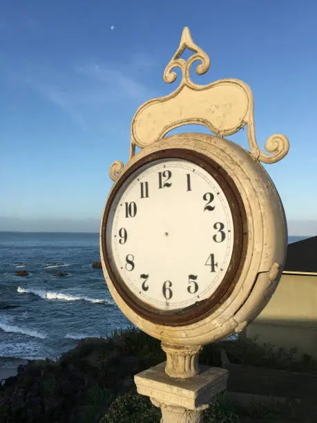 Large exterior clock by ocean with no hands, sea and clear blue sky in background.