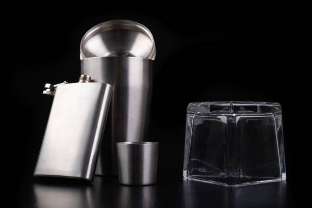 istock Metal shaker, hip flask and inverted vodka glass. Bartender accessories on the table. Black background. 1175996448