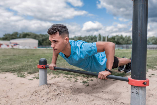 male athlete is pushing up from horizontal bar, training is powerful, in summer in city, in afternoon in fresh air. Youth lifestyle, active modern guy athlete. Background grass sand sky. stock photo