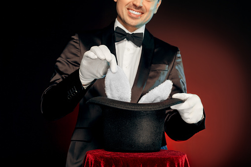 Professional magician wearing suit and gloves standing isolated on black and red background making trick taking rabbit from top hat on table close-up smiling playful.