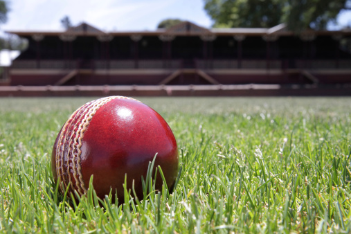 Shiny new cricket ball on grass in front of grand stand.