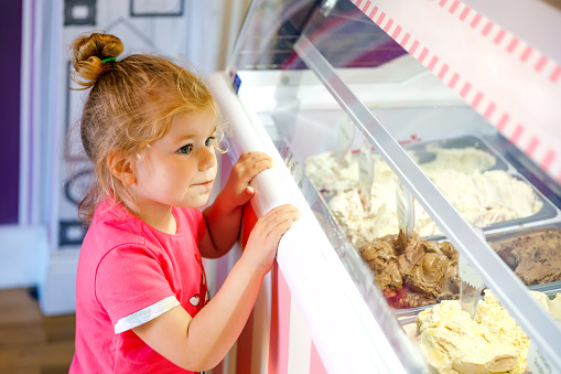 Cute little toddler girl choosing and buying ice cream in a cafe. Happy baby child looking at different sorts of icecream. Sweet home made dessert