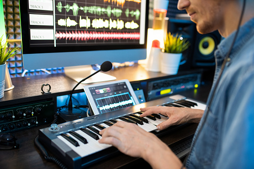 Young musician sitting by workplace in front of mike and pressing keys of piano keyboard while recording new music in studio