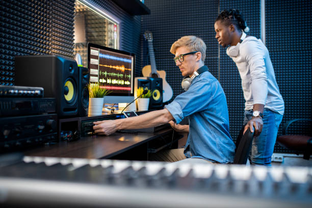 Young casual man adding volume while recording music with his African colleague Young casual man adding volume while recording music with his African colleague in contemporary studio sound technician stock pictures, royalty-free photos & images