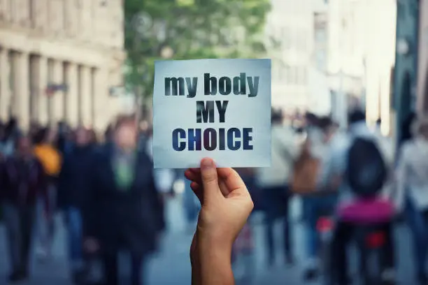 Photo of Feminist hands holding a protest banner with the message my body my choice over a crowded street. Human rights concept against fetus law and reproductive justice. Stop discrimination and injustice