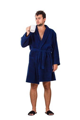 Full length portrait of sleepy young man messy hair, wears blue bathrobe holding a cup of tea in his hand, looking confused isolated on white background.