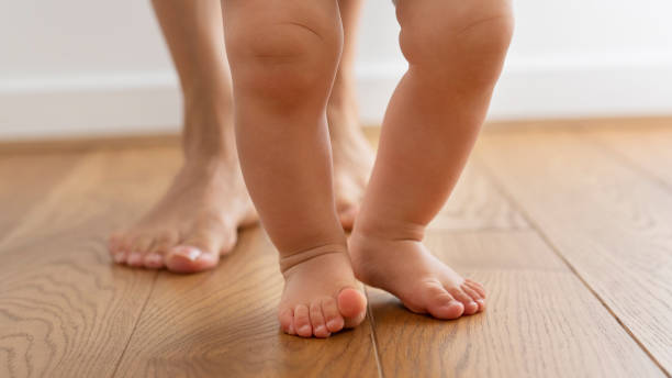 primo piano dei piedi mentre la madre aiuta il figlio piccolo a fare i primi passi a casa - barefoot foto e immagini stock