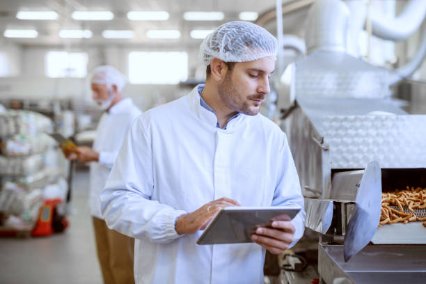 joven supervisor serio del cáucaso que evalúa la calidad de los alimentos en la planta de alimentos mientras se sostiene la tableta. el hombre está vestido con uniforme blanco y tiene red para el cabello. - sales manager fotografías e imágenes de stock