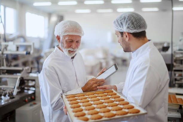 Young Caucasian employee holding tray with fresh cookies while supervisor evaluating quality and holding tablet. Both are dressed in sterile white uniforms and having hairnets. Food plant interior. joven empleado caucásico sosteniendo bandeja con galletas frescas mientras supervisor evalúa la calidad y la tableta de retención. ambos están vestidos con uniformes blancos estériles y con redes para el cabello. interior de la planta de alimentos.