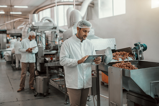 Joven supervisor serio del Cáucaso que evalúa la calidad de los alimentos en la planta de alimentos mientras se sostiene la tableta. El hombre está vestido con uniforme blanco y tiene red para el cabello. photo