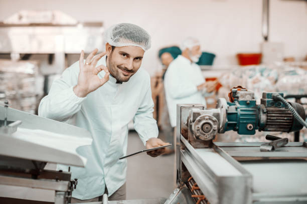 joven supervisor sonriente caucásico evaluando la calidad de los alimentos en la planta de alimentos mientras sostiene la tableta y muestra bien la señal. el hombre está vestido con uniforme blanco y tiene red para el cabello. - sales manager fotografías e imágenes de stock