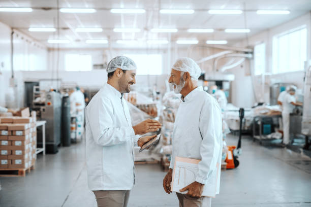istock Two supervisors talking about quality of food. Younger one holding tablet while older one holding folder with charts. Both are dressed in sterile uniforms and having hairnets. Food plant interior. 1176094780