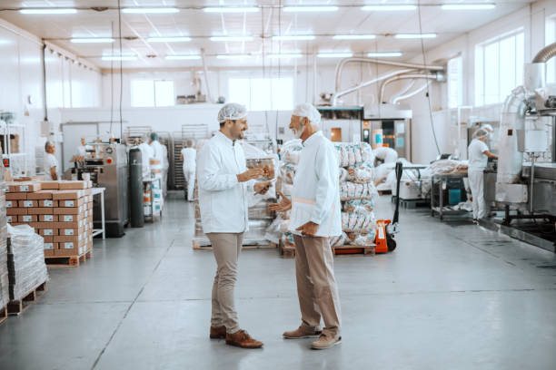 Two supervisors talking about quality of food. Younger one holding tablet while older one holding folder with charts. Both are dressed in sterile uniforms and having hairnets. Food plant interior.