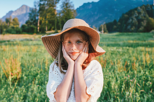 Beautiful romantic preteen girl in straw hat against the background of mountains, golden hour