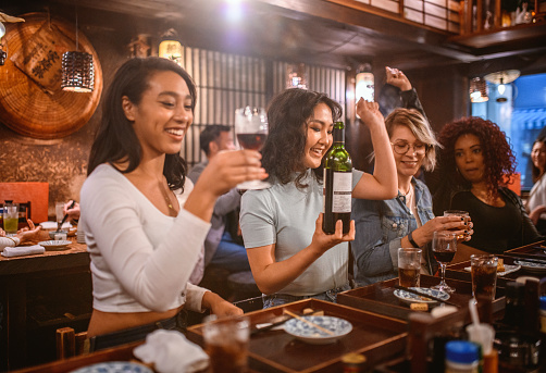 Group of laughing multi-ethnic female friends sitting at sushi bar in Tokyo izakaya and enjoying drinks.