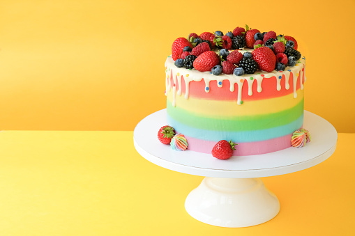 Cake on birthday with colorful rainbow cream on a yellow background decorated with berries, colorful sprinkles, poured with chocolate.