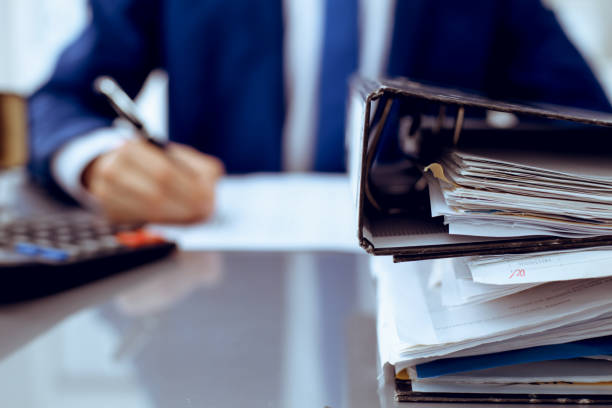istock Binders with papers waiting to be processed with businessman or bookkeeper back in blur. Accounting planning budget. Audit, insurance and business concept 1176148398