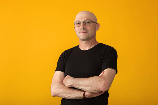 Studio portrait of a 50 year old man with a shaved head in a black T-shirt and glasses on a yellow background stock photo