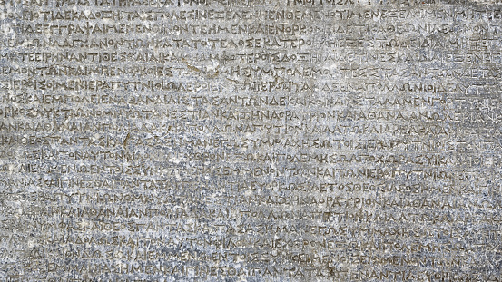 Ancient Greek writing on rock for background. Antique inscription carved on stone. Old script text close-up. Gray wall with historic letters. Vintage texture with words from past civilization.