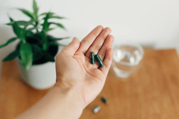 Dietary supplements. Hand holding spirulina capsules above glass of water on wooden table. Morning vitamin nutrient pill.  Health support and treatment. Biologically active additives Dietary supplements. Hand holding spirulina capsules above glass of water on wooden table. Morning vitamin nutrient pill.  Health support and treatment. Biologically active additives spirulina stock pictures, royalty-free photos & images