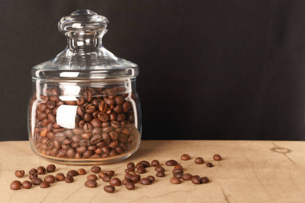 Roasted coffee beans in glass jar with lid. On light wooden surface, with dark background. With copy-space for text or logo.