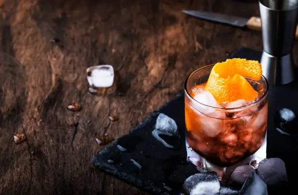 Red cocktail with dry vermouth, bitter, soda, orange zest and ice, wooden bar counter background, bar tools, selective focus
