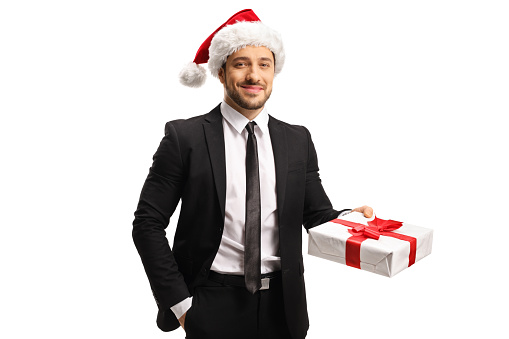 Young handsome man in a suit with a christmas hat holding a present isolated on white background