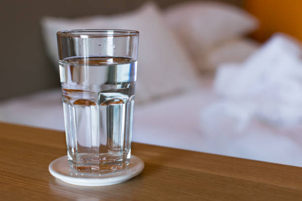 Glass of water in the morning. A glass of water sitting on a table next to a white bed in the morning, wake up routine. fast water stock pictures, royalty-free photos & images