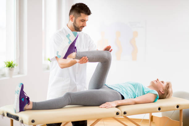 donna di mezza età con infortunio al ginocchio sdraiata sul tavolo della fisioterapia durante la sessione con giovane bel medico - physical therapy back table patient foto e immagini stock