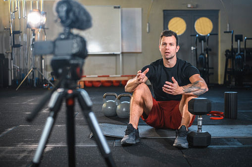 Caucasian male fitness instructor sitting on exercise mat at gym and making vlog explaining how to workout with kettlebells and dumbbells.