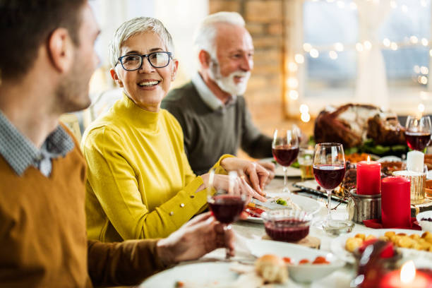 Happy senior woman eating Thanksgiving dinner with her family at dining table. Happy mature woman having Thanksgiving meal with her husband and talking to her adult son at dining table. multi generation family christmas stock pictures, royalty-free photos & images