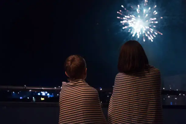 Mother and son looking at New Year celebration fireworks in night sky warmly wrapped in striped plaids standing on home balcony.