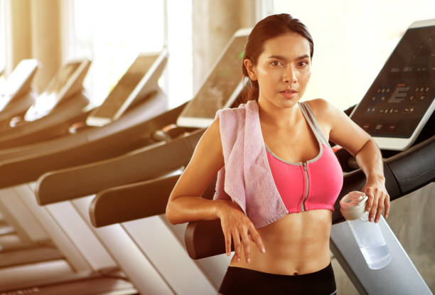 istock Beautiful Asian young woman tired taking a break from running or exercise standing on treadmill machine drinking water and towel sweat in fitness gym healthy .girl in sportswear workout rest 1176341944