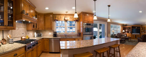 Wide panoramic photo of an updated kitchen and great room with a center island, bar stools, stainless steel appliances, granite countertops