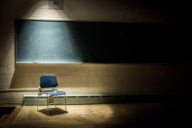 una silla escolar vacía en un aula oscura y sombría - frente a una pizarra con un solo haz de luz en la cabeza - vacío fotografías e imágenes de stock